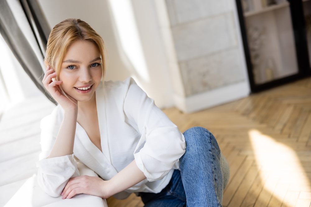 A woman smiling while seated indoors, wearing a white blouse and jeans - Seattle Dentist A woman smiling while seated indoors, wearing a white blouse and jeans - Seattle Dentist