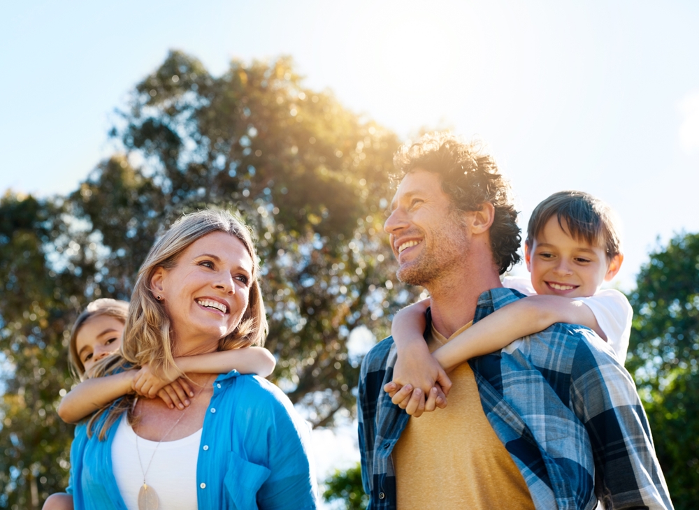 A smiling family walking outdoors, with two children riding on their parents' backs - Seattle Dentist A smiling family walking outdoors, with two children riding on their parents' backs - Seattle Dentist