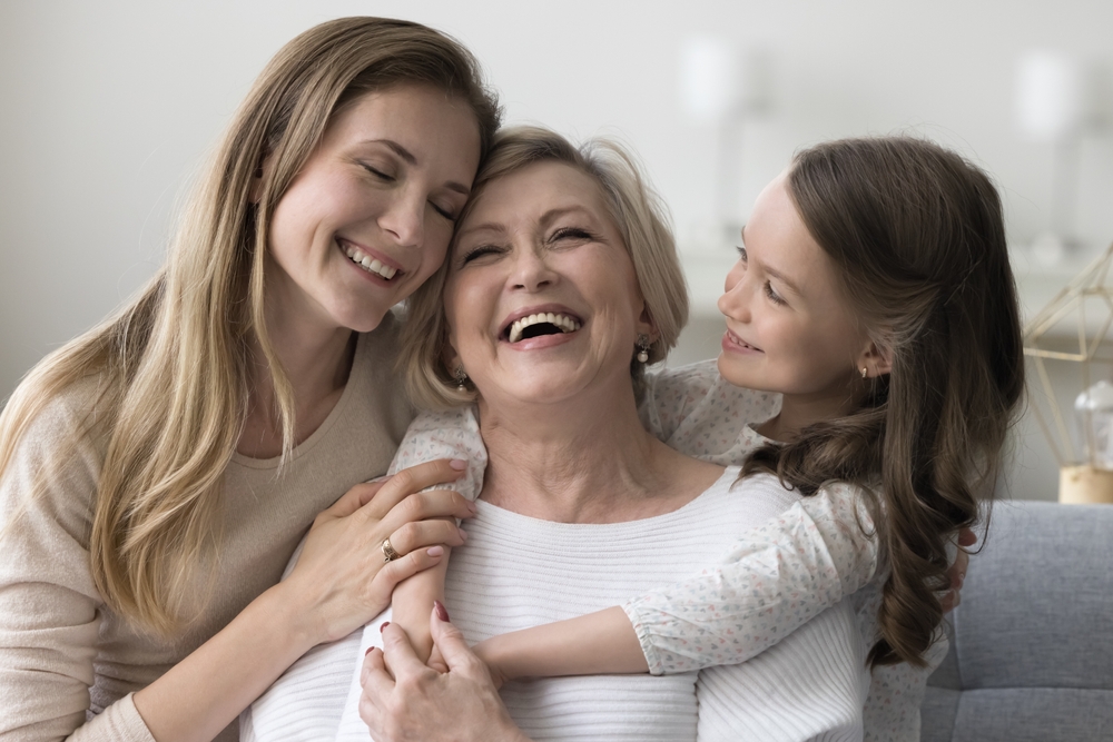 A grandmother, daughter, and granddaughter share a joyful embrace, all smiling together - Seattle Dentist A grandmother, daughter, and granddaughter share a joyful embrace, all smiling together - Seattle Dentist
