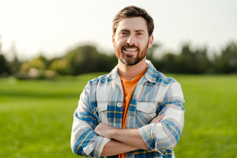 A man smiling confidently outdoors in a plaid shirt with arms crossed - Seattle Dentist A man smiling confidently outdoors in a plaid shirt with arms crossed - Seattle Dentist